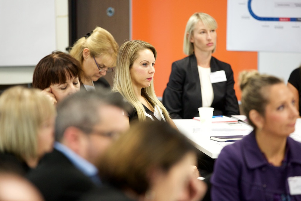 A group of people sit at tables looking to the front of the room where a presenter is 