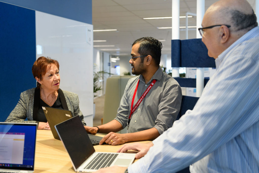 3 people stand at a table together talking with computers in front of them