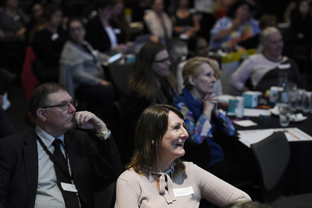 Forum attendees sit at tables and look away from camera towards speakers