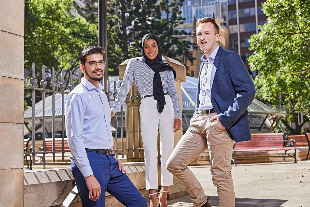 Three graduates (male, female, male) standing outside on stairs and smiling at camera