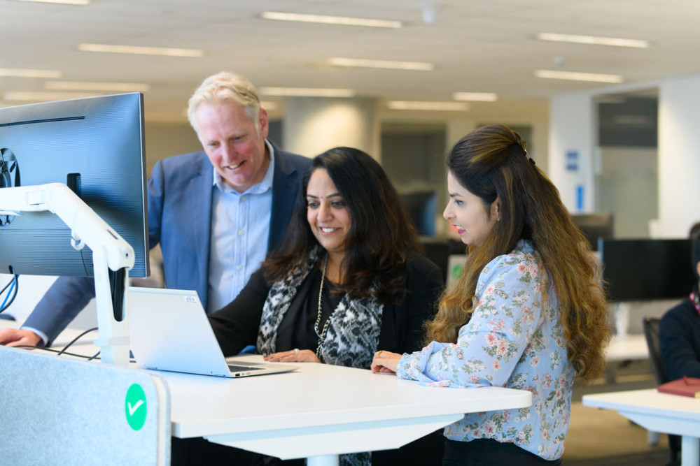 A man and 2 women standing while looking at a laptop on a desk