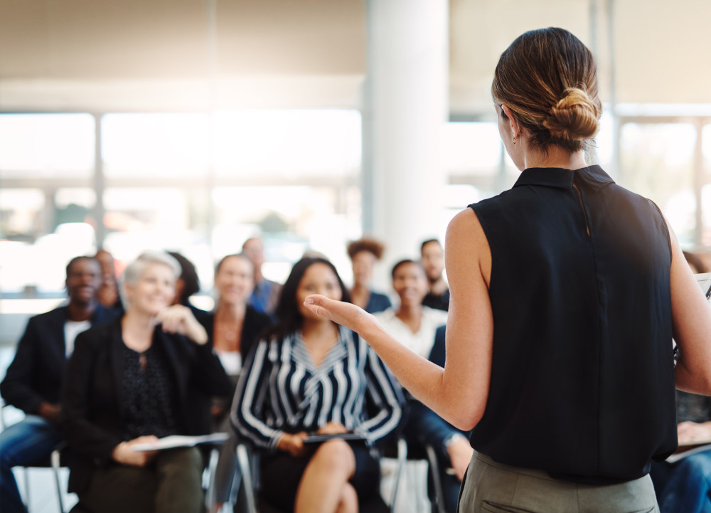 A woman standing in front of a seated crowd speaking