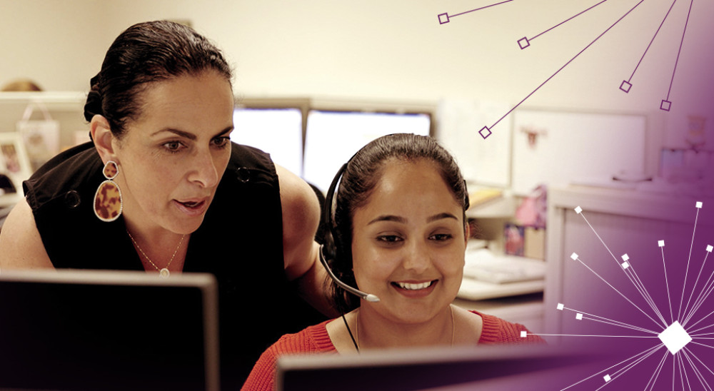 A woman stands next to another woman working on a computer